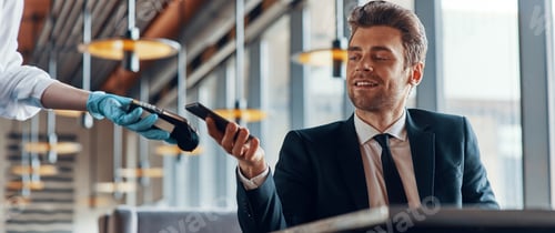 Preview: Handsome young smiling man in full suit making a contactless payment while sitting in the restaurant