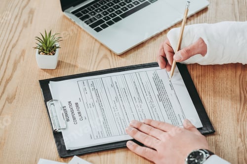 Preview: cropped view of man with broken arm in gypsum signing compensation form on table in office,