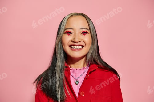 Preview: Valentines day concept, cheerful asian woman with heart shaped eye makeup posing on pink backdrop