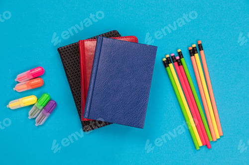 Preview: Colorful textliners and crayons and three dayplanners in leather covers are lying on pale-blue