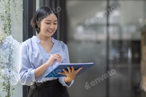 Preview: Boring young business Asian woman holding document file and looking at camera stand on a workplace o