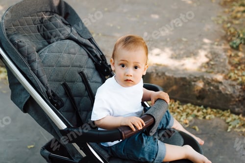 Preview: Portrait of a funny little child sitting in a stroller and looking at the camera, outdoors. Kid in a
