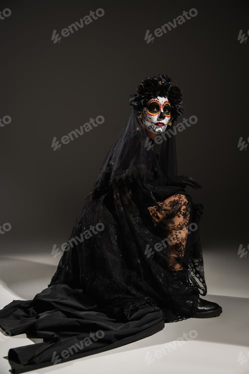 Preview: full length of woman with sugar skull makeup wearing black dress with lace veil posing on dark
