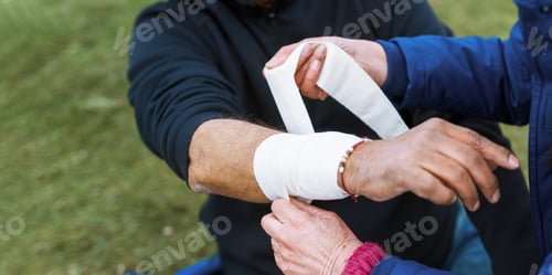 Preview: Close-up of a person bandaging another's arm outdoors.