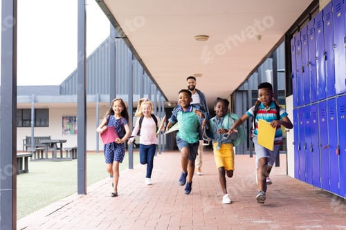 Preview: Happy, diverse male teacher with children running in elementary school corridor, copy space