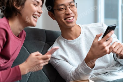 Preview: Photo of joyful multinational couple laughing and using smartphones