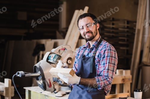 Preview: Handsome carpenter in protective glasses is looking at camera and smiling while standing near his