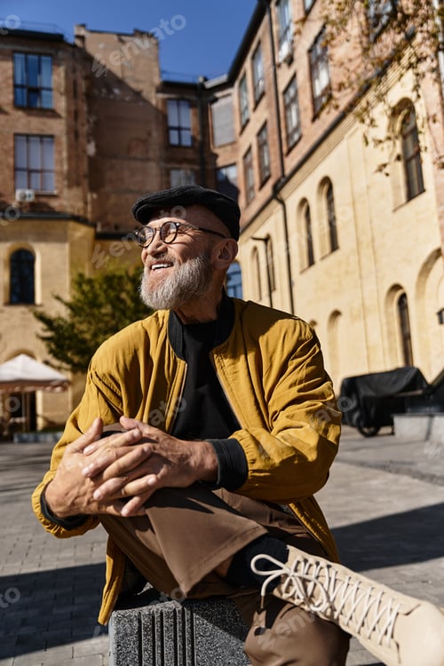 Preview: A handsome senior man sits comfortably in a chic city square, dressed in a trendy jacket and pants.