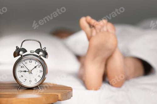 Preview: Feet of caucasian millennial lady lies on bed under white blanket, sleep at morning, focus on alarm