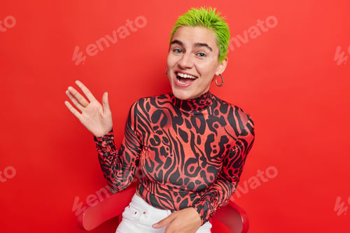 Preview: Positive female teenager with unique appearance has trendy green hairstyle waves palm greets you has