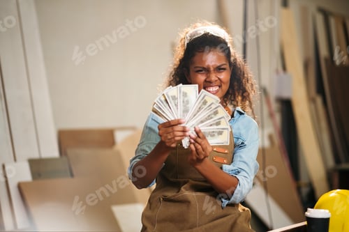 Preview: Young attractive black woman carpenter holding banknote from selling wood furniture in wood workshop