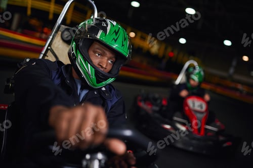 Preview: african american man in helmet driving go kart on indoor circuit near friend on blurred backdrop