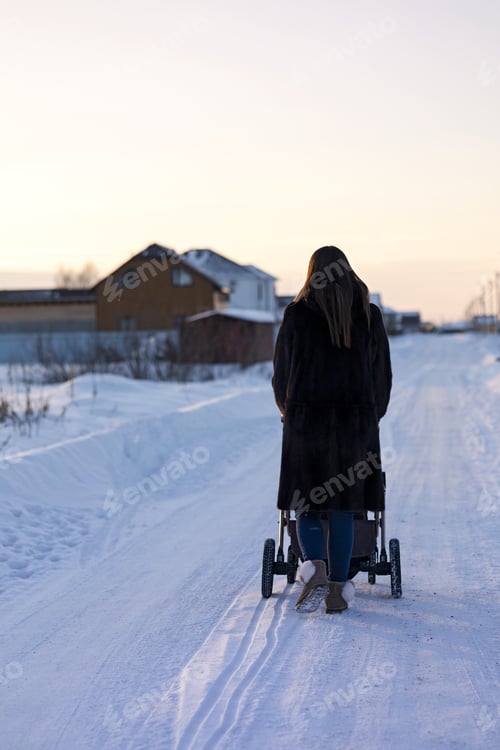 Preview: Young beautiful mother walking with her baby in a stroller in winter in snowy countryside