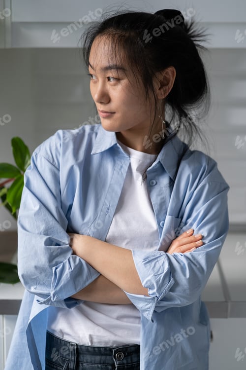 Preview: Pensive young Asian girl standing at home with arms crossed looking away thinking of future