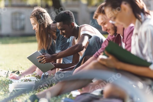 Preview: side view of young multiethnic students studying and reading books while sitting on grass