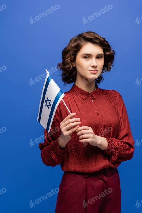 Preview: young and serious language teacher with flag of Israel looking at camera isolated on blue