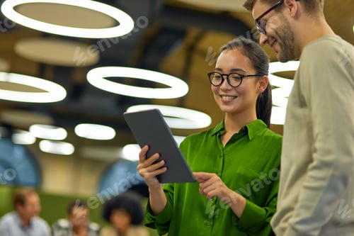 Preview: Young cheerful asian woman showing something on digital tablet to her male colleague while standing
