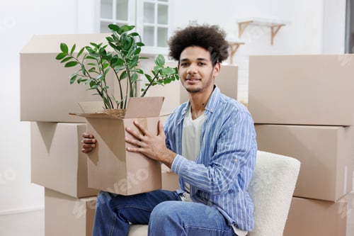 Preview: happy young African American man moves into new home. person is sitting among cardboard boxes