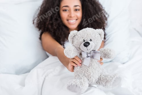 Preview: selective focus of african american girl laying in bed and showing teddy bear at home