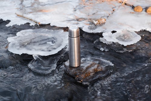 Preview: Thermos standing in a winter stream. Vacuum steel flask for hiking. Travel concept, warming drink in