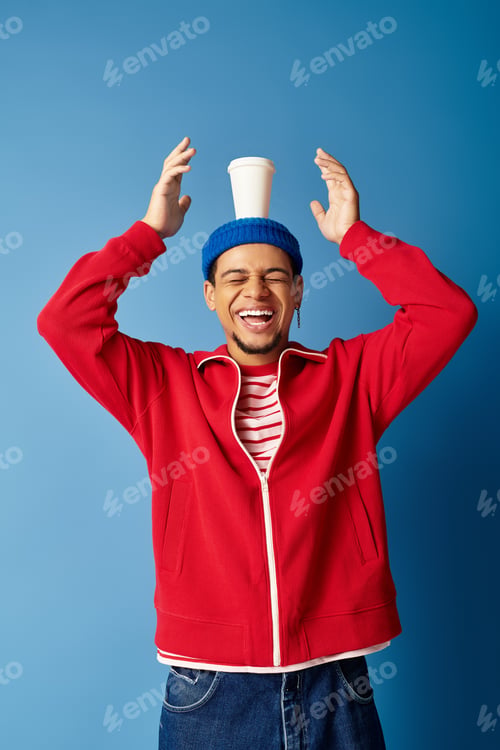 Preview: Stylish young man with beanie and playful spirit against a vibrant blue backdrop
