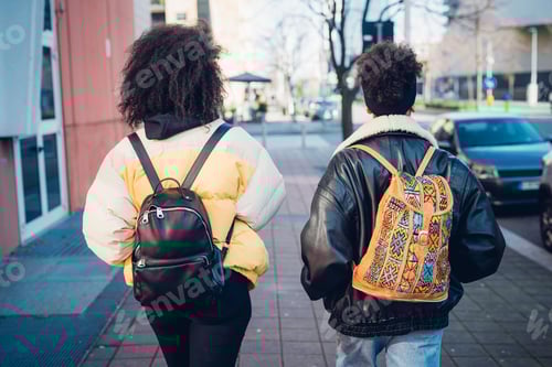 Preview: Two young women carrying backpacks strolling on urban sidewalk, rear view