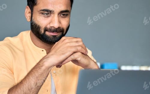 Preview: Thoughtful indian businessman looking at laptop watching business webinar.
