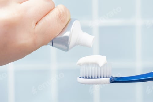 Preview: Female hand squeezing toothpaste onto toothbrush against blue tile background.