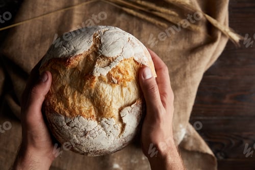 Preview: cropped shot of male baker holding bread over wooden table with wheat and sackcloth
