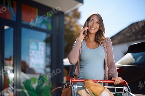 Preview: Happy pregnant woman talking on mobile phone in front of a supermarket.