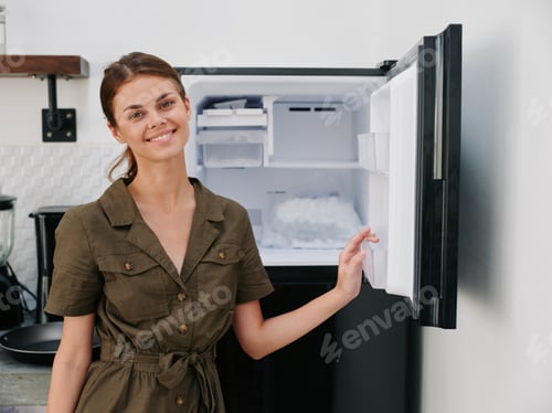 Preview: Woman smiling with teeth looking into camera in kitchen at home opened freezer empty with ice inside