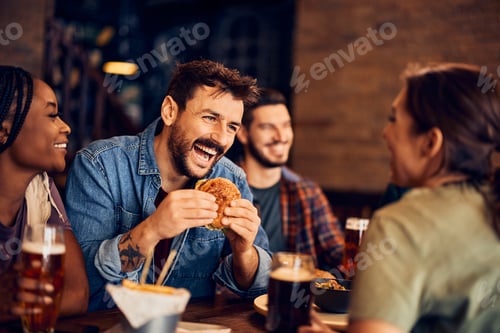 Preview: Happy man eating burger while talking to his friends in pub.