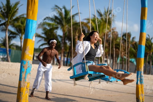Preview: Happy interracial couple enjoying a fun beach day, swinging under palm trees on a tropical vacation