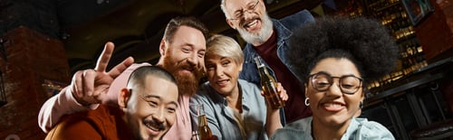 Preview: cheerful bearded man showing victory sign near multiethnic friends with beer bottles in bar, banner