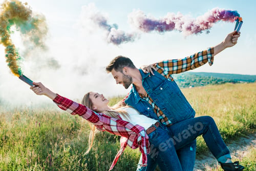 Preview: Side View of Man With Colorful Smoke Bombs Holding Girlfriend Back in Rural Field