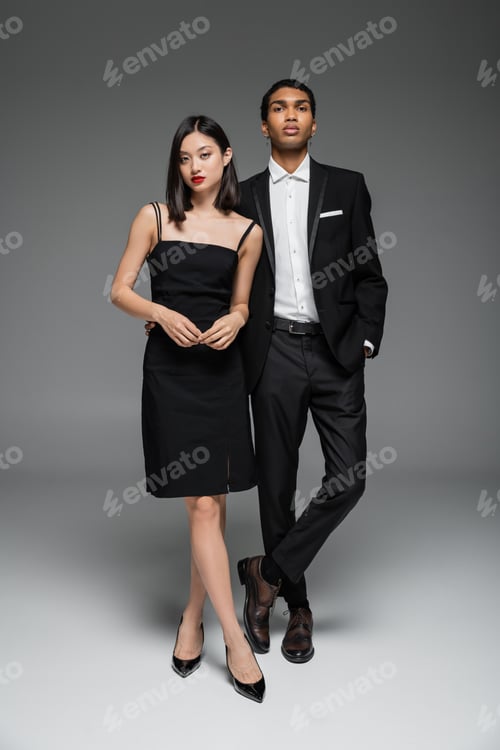 Preview: full length of asian woman in black dress and african american man in stylish suit standing on grey