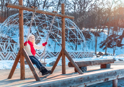 Preview: Happy middle aged blonde woman in white sweater, red scarf and gloves riding a swing at winter day.