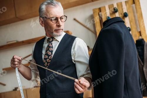 Preview: senior tailor holding tape measure and looking at jacket at sewing workshop