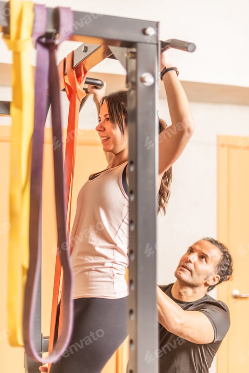 Preview: Cropped view of a athlete woman doing pull ups exercise with bar with help of trainer in gym