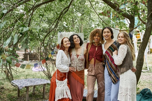 Preview: positive and multiethnic women in boho outfit looking at camera in retreat center