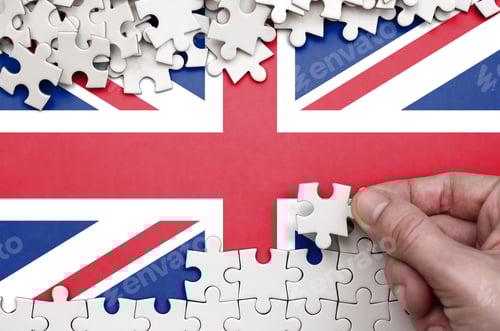 Preview: Great britain flag is depicted on a table on which the human hand folds a puzzle of white color.