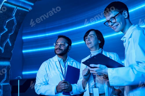 Preview: young intern with clipboard smiling near multiethnic colleagues in futuristic science center