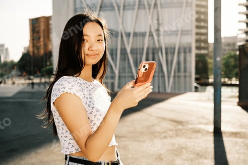 Preview: Young Asian woman looking at camera while using her mobile phone outdoors on the street.
