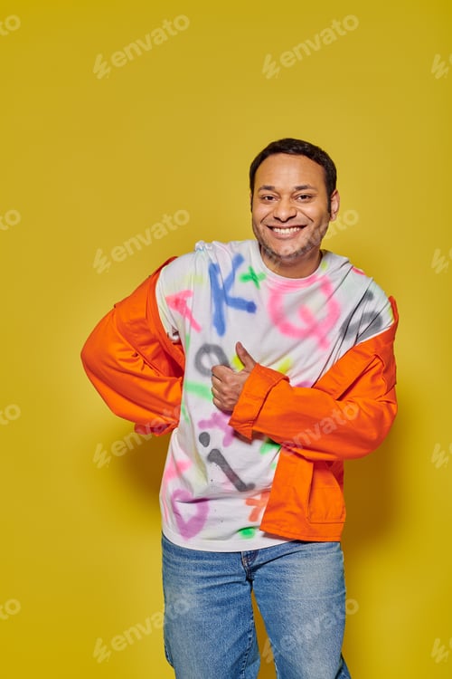 Preview: positive indian man in orange jacket and diy t-shirt showing thumb up on yellow background