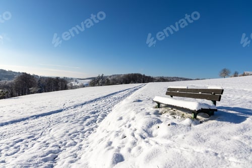 Preview: Empty bench covered in snow in a field on a clear winter sunny day