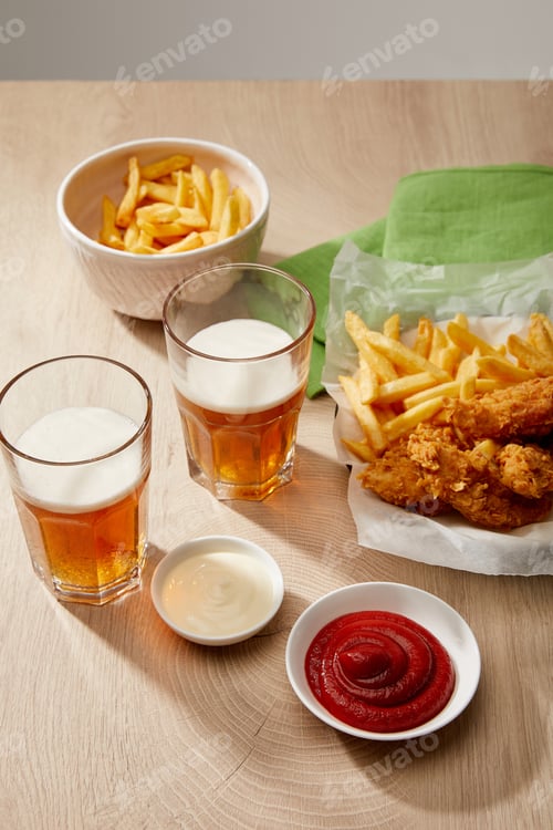 Preview: glasses of beer, chicken nuggets with french fries, ketchup and mayonnaise on wooden table on grey