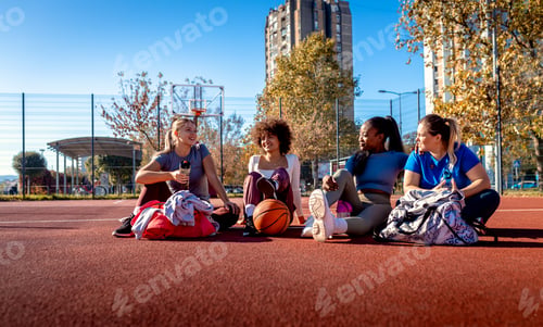 Preview: Diverse group of young woman sitting on court resting afrer playing basketball outdoors.