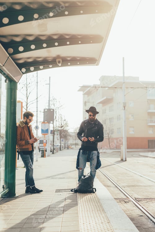 Preview: Two young male hipsters looking at smartphones while waiting at city tram station