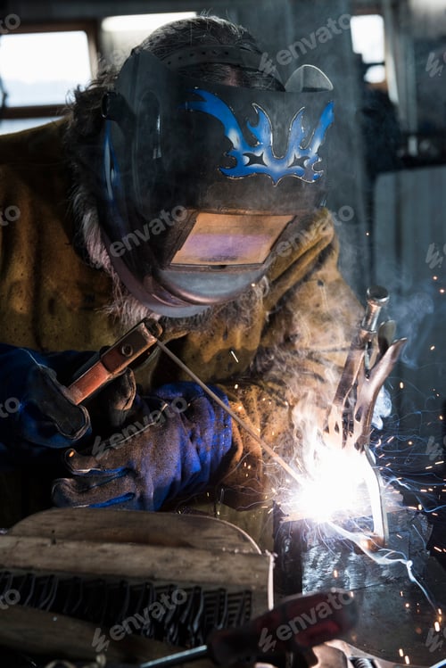 Preview: Blacksmith in welding mask welding metal in workshop