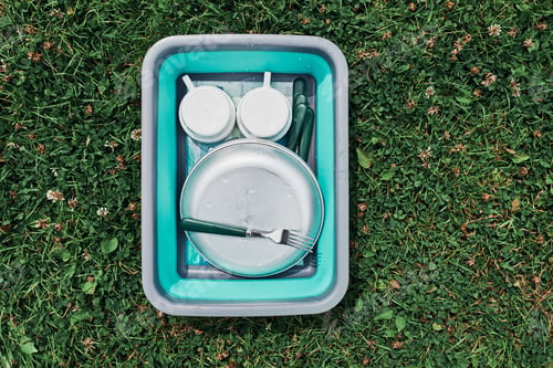 Preview: Washing up bowl filled with the washed outdoor dishes, plates, cups and cutlery put on grass